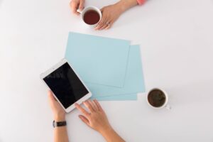 two pairs of hands on desk with one holding coffee and ther other holding a tablet computer